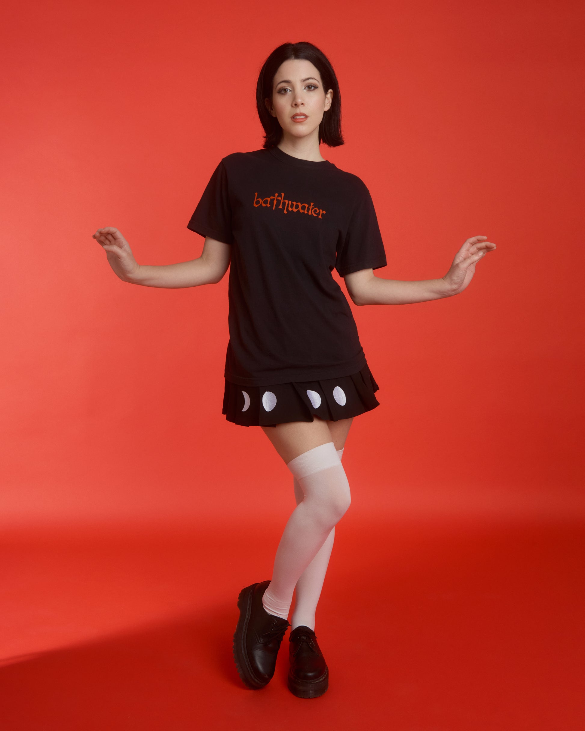 A woman with short black hair wearing a black t-shirt that says "bathwater" in red ink, posing in front of a bright red background.